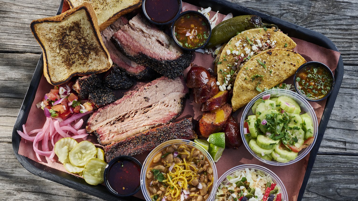 Vaqueros Texas BBQ tray with sliced brisket, ribs, toast, sides, pickles, and a cold beer